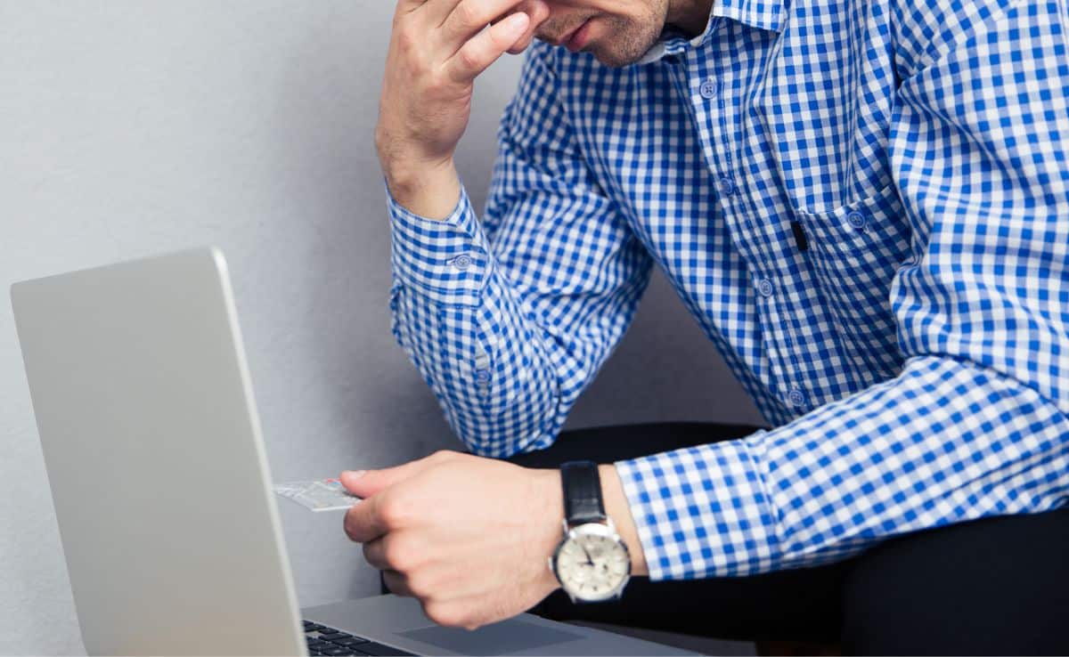 A depressed man holding credit card over gray background wearing a watch and blue checkered long sleeve shirt