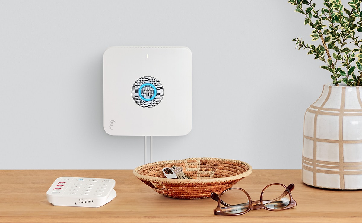 Amazon Ring base station hangs on wall with Ring keypad sitting on credenza in foreground. Keypad sits next to basket with keys, glasses, and potted plant.