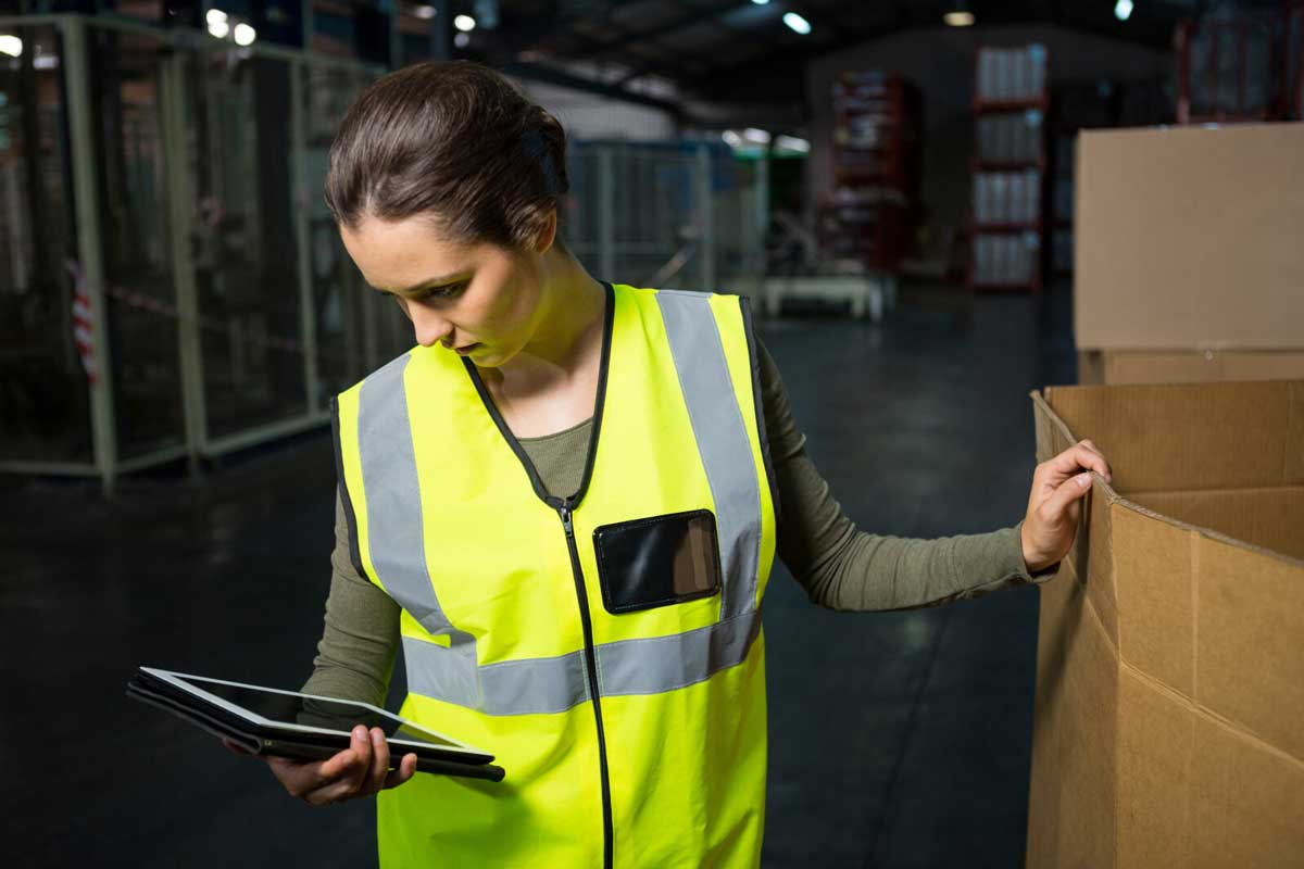 Delivery lady in reflective uniform checking tablet, with her other hand resting on a large cardboard box.