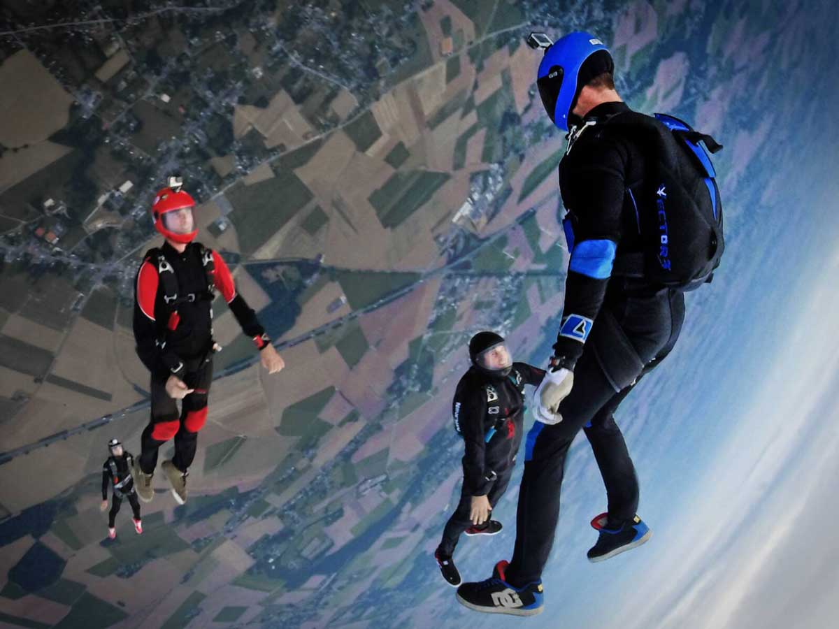 Four people skydiving, camera at an angle, with the ground viewable far below them