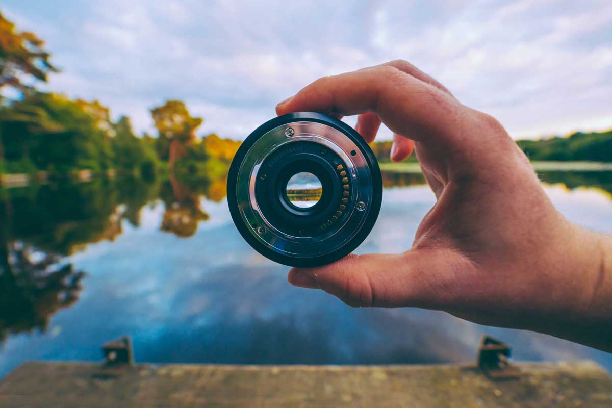 Hand holding a camera lens with lake and trees in background