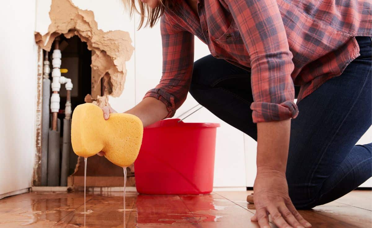 Worried woman mopping up water on the floor from a burst pipe with sponge with hole in the wall in the background