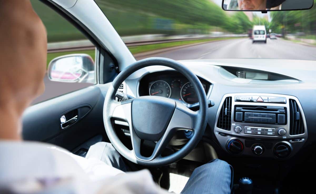 A man sitting in the driver's seat of a self driving car with hands off the steering wheel wearing suit with a blurry street scene