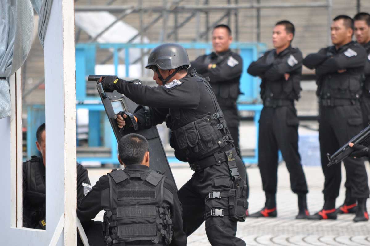 Police in incidence response training. An officer moving forward wearing shield and helmet, weapon drawn. Four offices with arms crossed looking on.