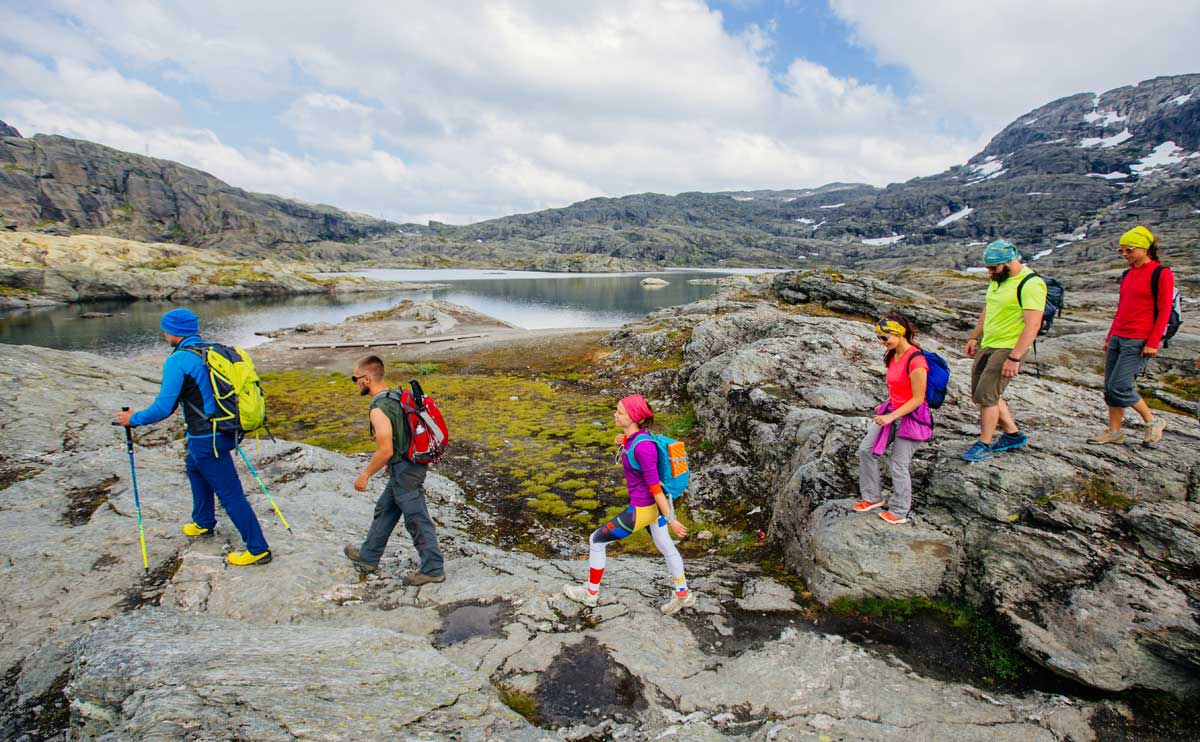 Group of six hikers men and woman walking with backpacks and trekking poles along water lake in mountain valley during Norway travel