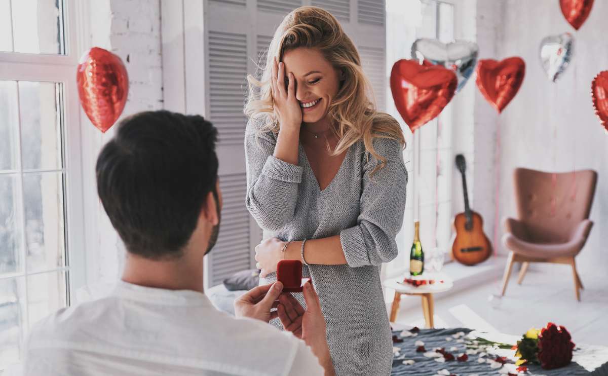 Surprised young woman covering face with hand and smiling while her boyfriend proposing her in the bedroom.
