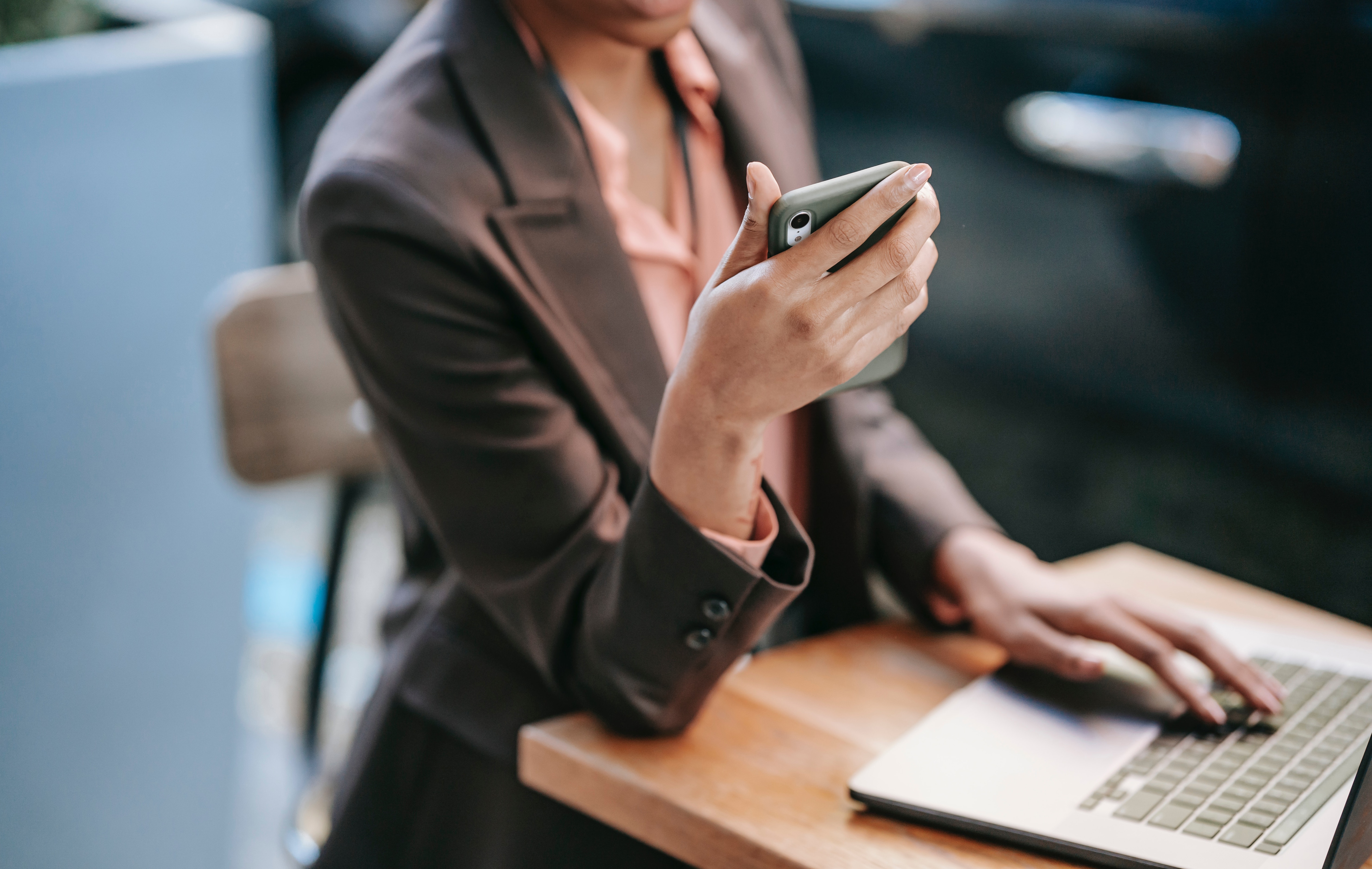 A woman in a business suit is checking her phone and working on a laptop as she sits outside at a table.