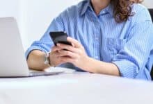Woman sitting at her desk in front of computer on cell phone.