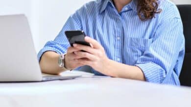 Woman sitting at her desk in front of computer on cell phone.