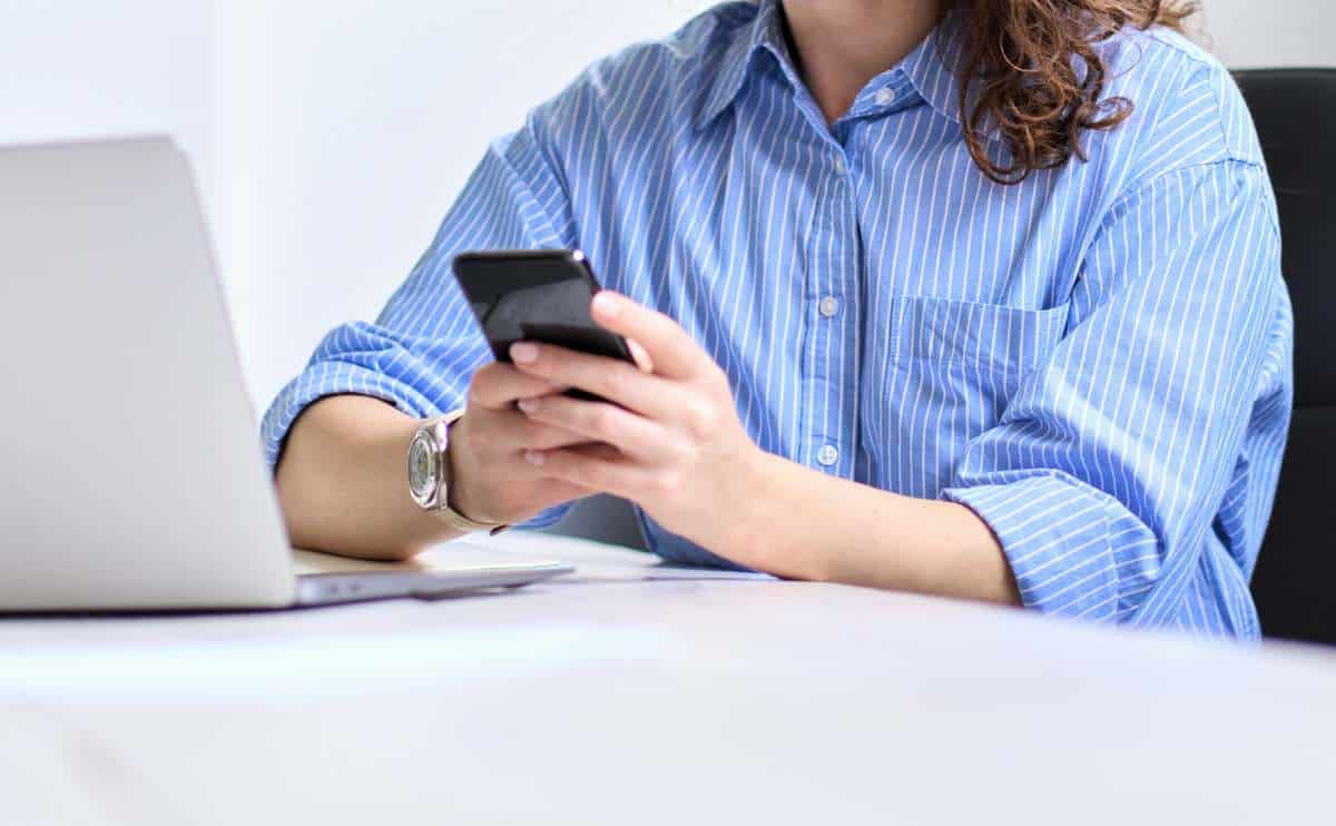 Woman sitting at her desk in front of computer on cell phone.