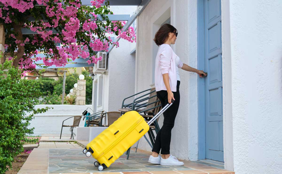 Young woman with yellow suitcase arriving at blue door of condo, surrounded by pink spring blooms.